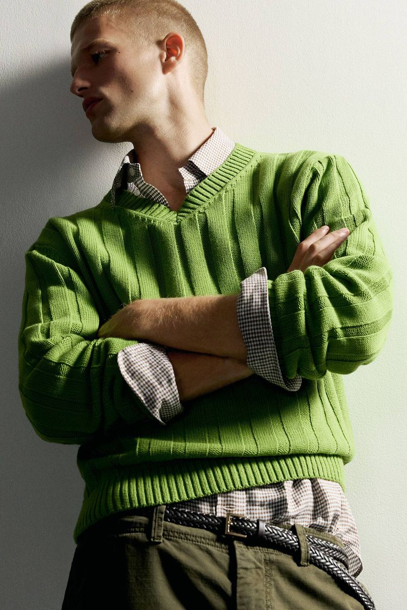 Model with short buzz cut wearing a bright green ribbed v-neck sweater and collared shirt with arms crossed against a white wall.