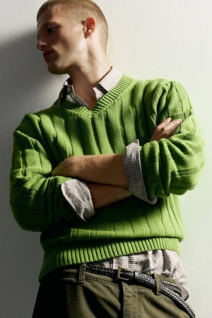 Model with short buzz cut wearing a bright green ribbed v-neck sweater and collared shirt with arms crossed against a white wall.
