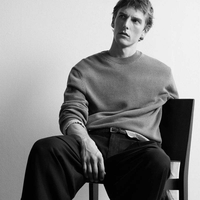 Man seated on a dark chair wearing a crewneck knit sweater and dark trousers in black and white studio lighting.