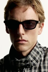 Close-up portrait of a man wearing dark sunglasses and a black and white plaid zippered jacket against a white background.