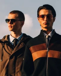 Two male models wearing aviator sunglasses, collared shirts, ties, and structured outerwear against a soft blue sky.