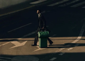 A man in a dark tailored coat walking across an asphalt street pulling a green suitcase and matching holdall bag.