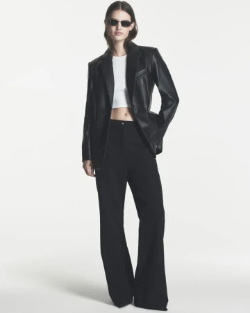 Model wearing a black leather blazer, white crop top, wide-leg black trousers, and dark sunglasses against a white background.