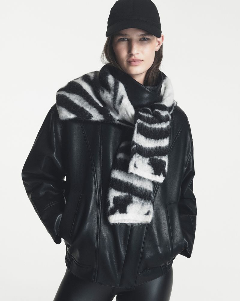 Model Emmi Freeman in a black leather jacket, black cap, and black-and-white zebra knit scarf against a white background.