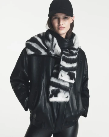 Model Emmi Freeman in a black leather jacket, black cap, and black-and-white zebra knit scarf against a white background.