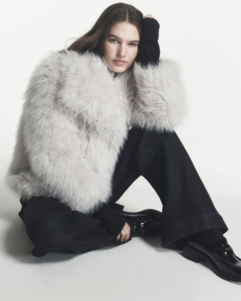 Model Emmi Freeman seated on the floor wearing a grey faux-fur coat, wide-leg trousers, and black patent loafers on a white backdrop.