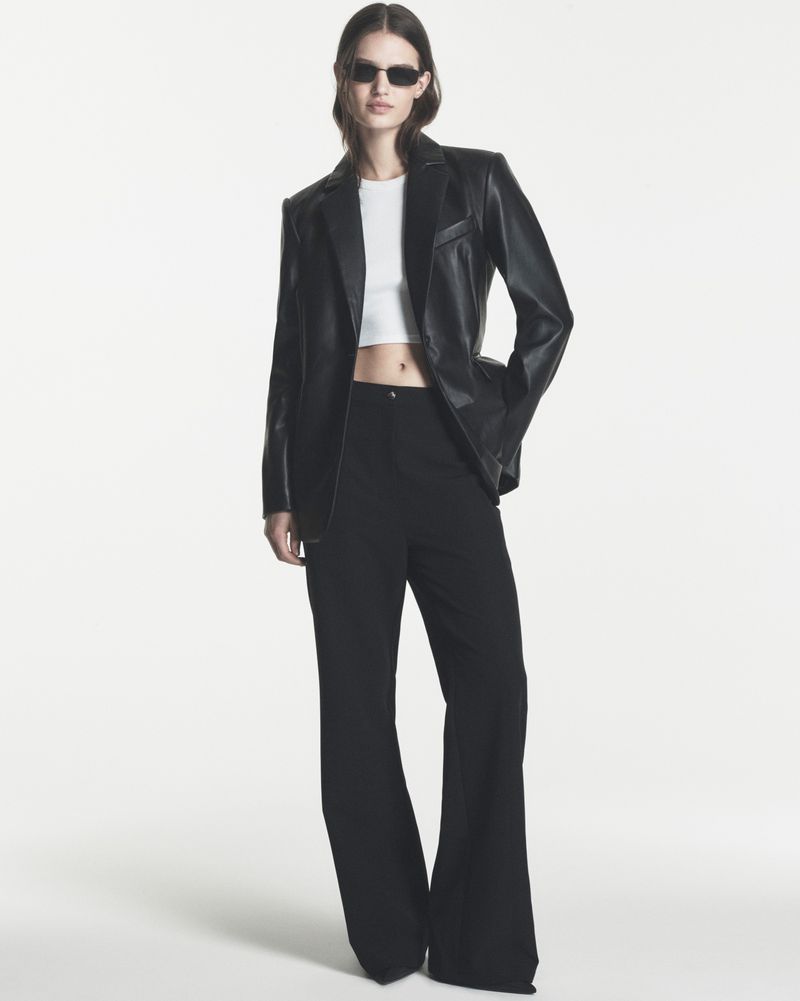 Model wearing a black leather blazer, white crop top, black wide-leg trousers, and sunglasses against a white backdrop.