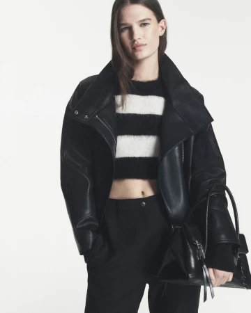 Model Emmi Freeman wearing an oversized black leather jacket, striped crop knit, and black trousers against a white seamless background.