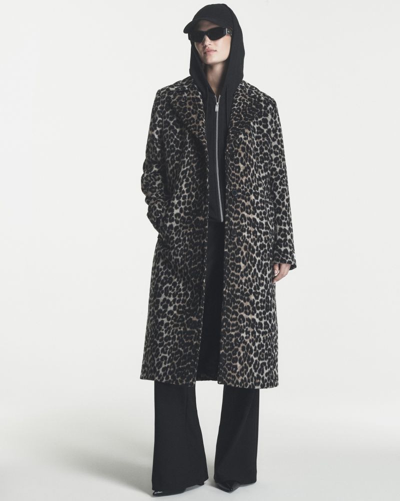 Model wearing a long leopard-print coat over a black hoodie and sunglasses against a white background.