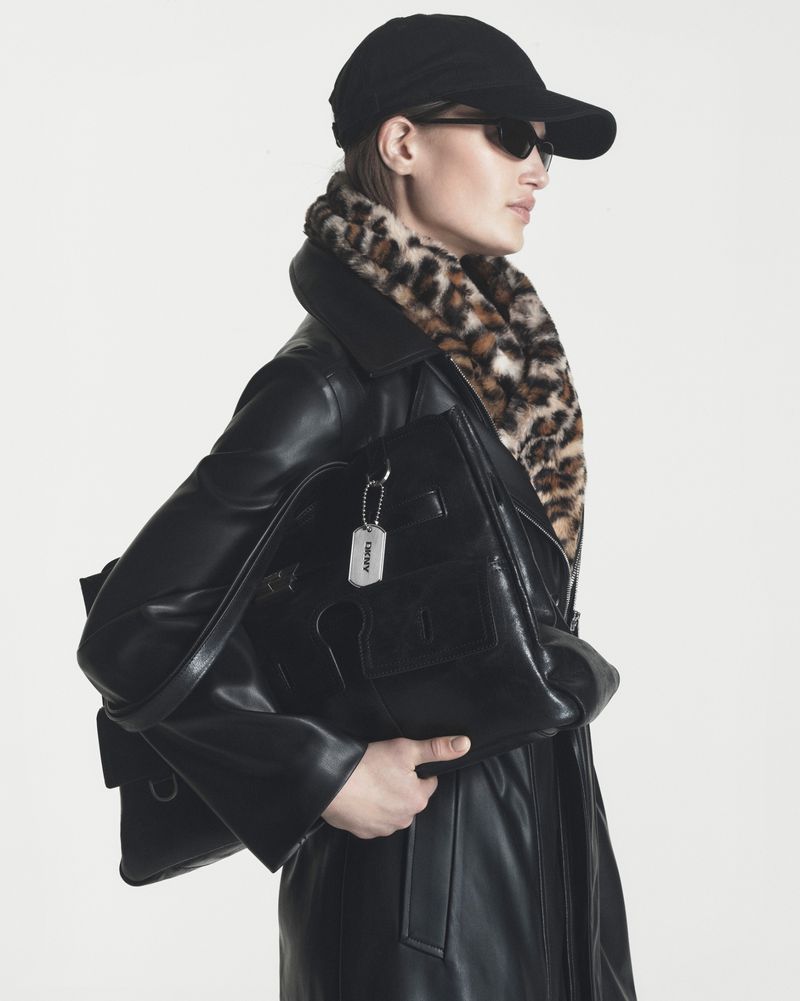 Model wearing a black leather coat, leopard print collar, cap, and sunglasses while holding a black leather tote bag against white.