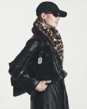 Model wearing a black leather coat, leopard print collar, cap, and sunglasses while holding a black leather tote bag against white.