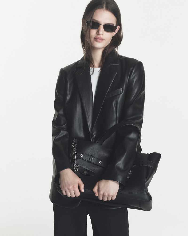 Model Emmi Freeman wears a black leather blazer and dark sunglasses while holding a black leather tote against a white backdrop.