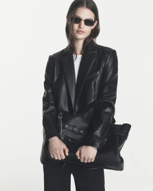 Model Emmi Freeman wears a black leather blazer and dark sunglasses while holding a black leather tote against a white backdrop.