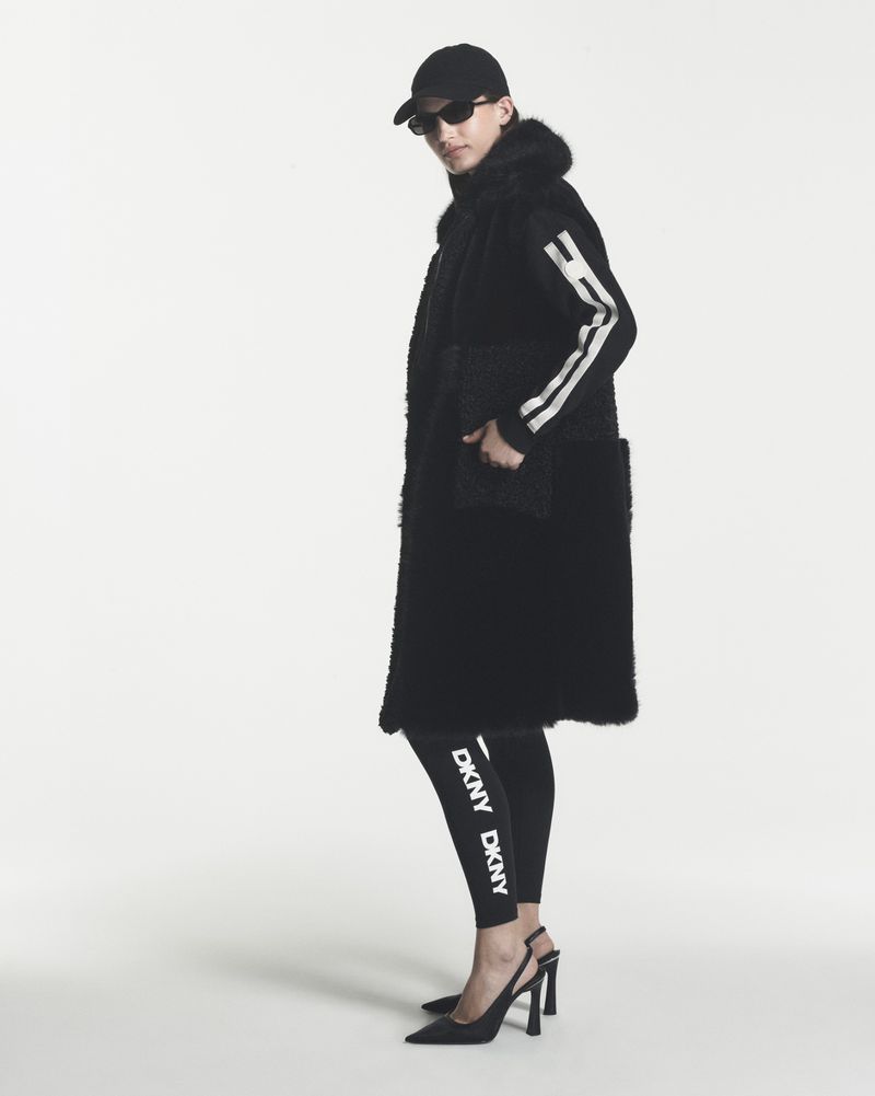Model wearing a long black shearling vest, DKNY branded leggings, sunglasses, and black slingback heels against a white backdrop.
