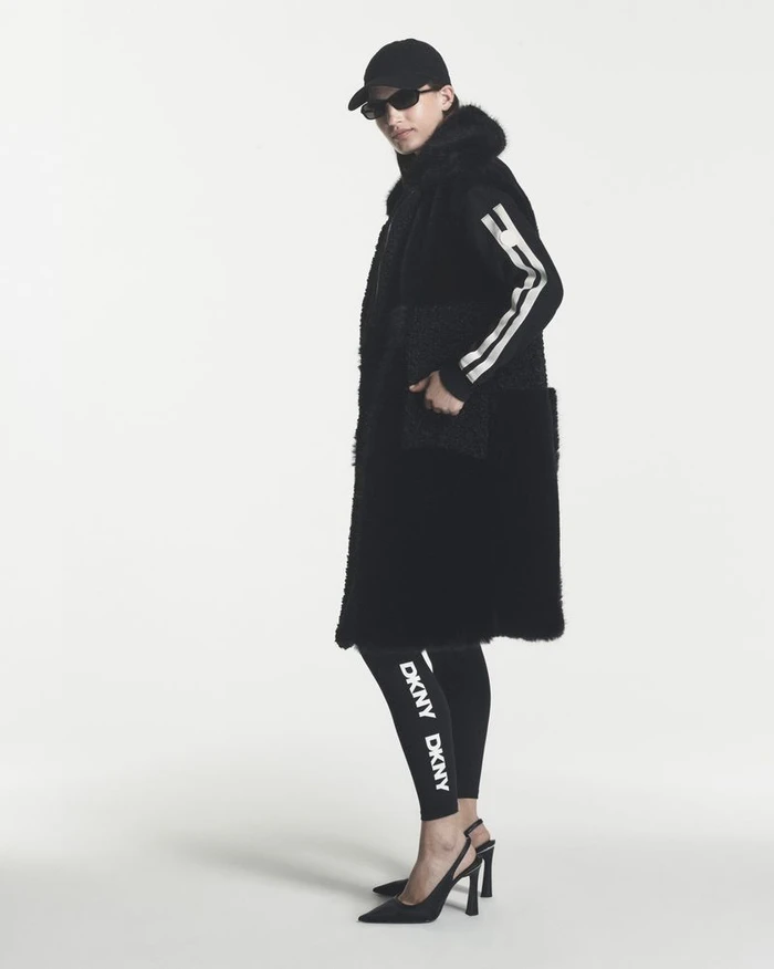 Model wearing a long black shearling vest, DKNY branded leggings, sunglasses, and black slingback heels against a white backdrop.