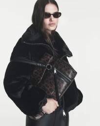 Model Emmi Freeman in black sunglasses, a black plush coat, and holding a leopard-print crossbody bag against a white studio background.