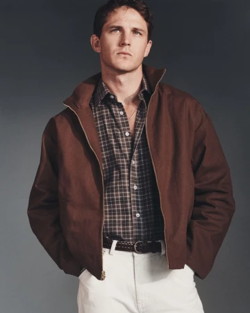 Model Tommy Hackett posing in a brown zip-up jacket, plaid collared shirt, and white trousers in front of a grey studio backdrop.
