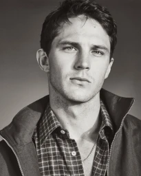 Monochrome portrait of model Tommy Hackett wearing a plaid shirt and open jacket against a neutral studio backdrop.
