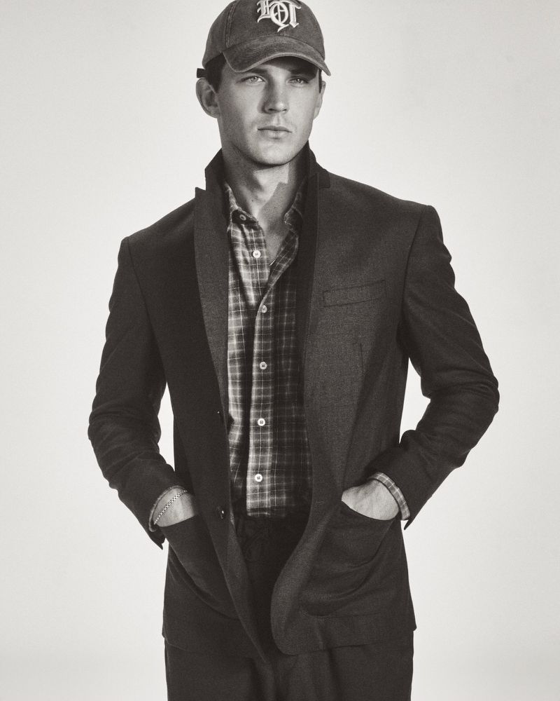 Model Tommy Hackett posing in a tailored dark blazer, plaid shirt, and embroidered baseball cap against a light seamless background.