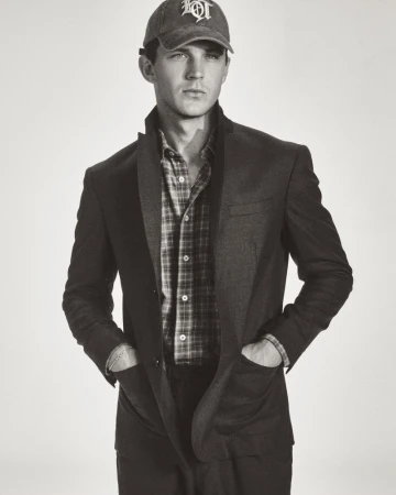 Model Tommy Hackett posing in a tailored dark blazer, plaid shirt, and embroidered baseball cap against a light seamless background.