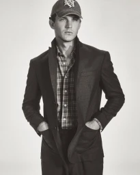 Model Tommy Hackett posing in a tailored dark blazer, plaid shirt, and embroidered baseball cap against a light seamless background.