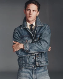 Model Tommy Hackett posing with arms crossed in a denim jacket, collared shirt, and tie against a studio grey backdrop.