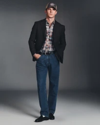 Tommy Hackett wearing a black blazer, plaid shirt, blue jeans, and a baseball cap against a grey studio backdrop.