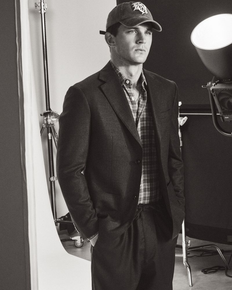 Model Tommy Hackett wearing a tailored blazer, plaid shirt, and baseball cap in a black and white studio portrait.