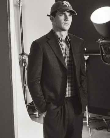 Model Tommy Hackett wearing a tailored blazer, plaid shirt, and baseball cap in a black and white studio portrait.