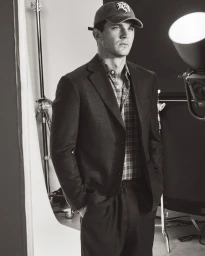 Model Tommy Hackett wearing a tailored blazer, plaid shirt, and baseball cap in a black and white studio portrait.