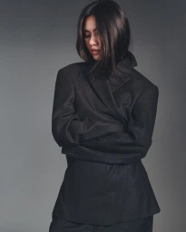 Model with long dark hair looking down with arms crossed, wearing a tailored black blazer against a grey backdrop.
