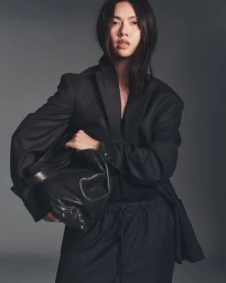 Model wearing an oversized black blazer and drawstring trousers, holding a slouchy black leather handbag against a grey backdrop.