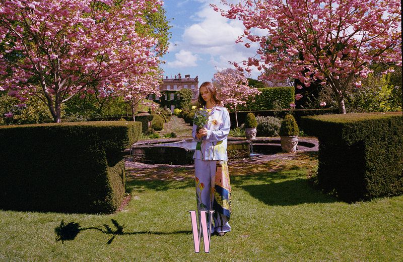 A young woman in a floral pajama set holding yellow flowers stands in a garden with pink cherry blossom trees and a house in background.