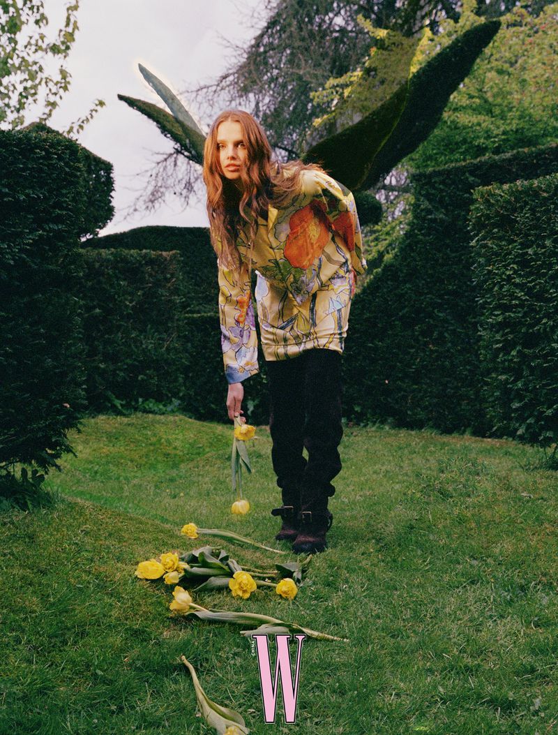 Adult woman in floral top and black pants kneeling in a garden with yellow tulips, set against a backdrop of green hedges.