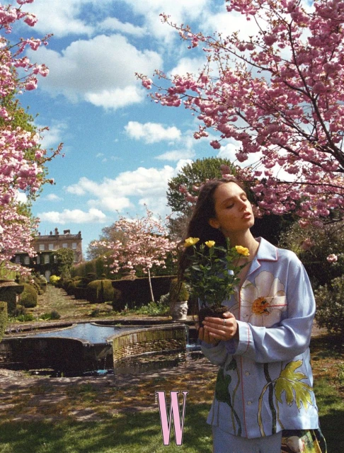 Woman in floral pajamas holds yellow roses in a garden setting with pink cherry blossoms and a historic building in the background.