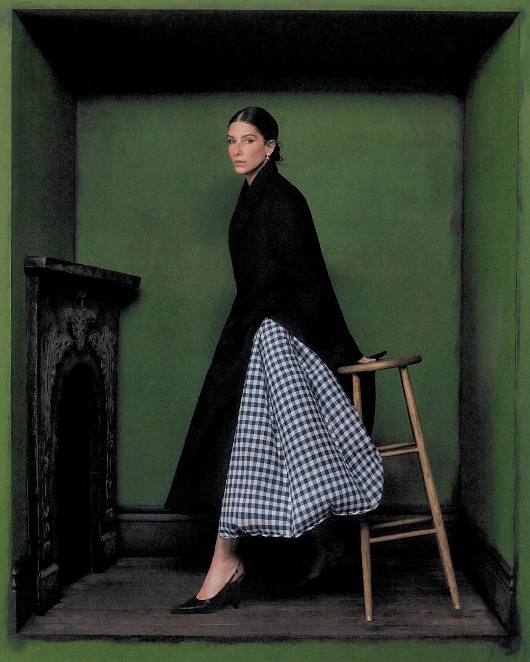 Sandra Bullock in a black coat and voluminous gingham skirt posing inside a green room set next to a wooden stool.