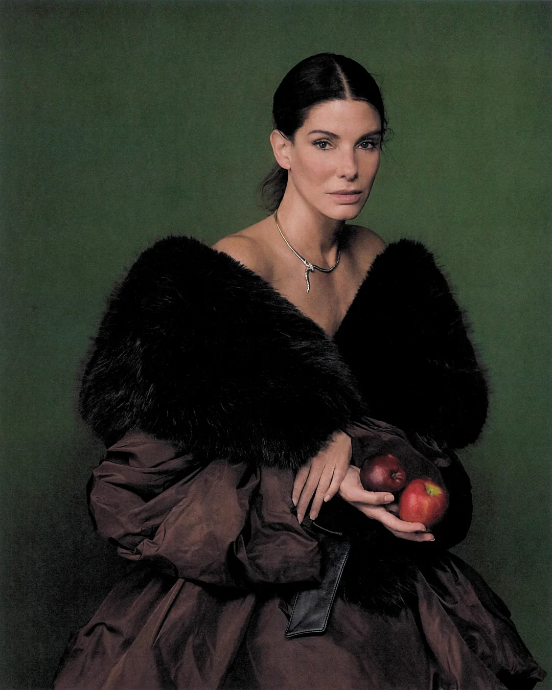 Sandra Bullock wearing a black fur stole and brown gown while holding two red apples against a dark green backdrop.
