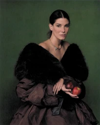 Sandra Bullock wearing a black fur stole and brown gown while holding two red apples against a dark green backdrop.