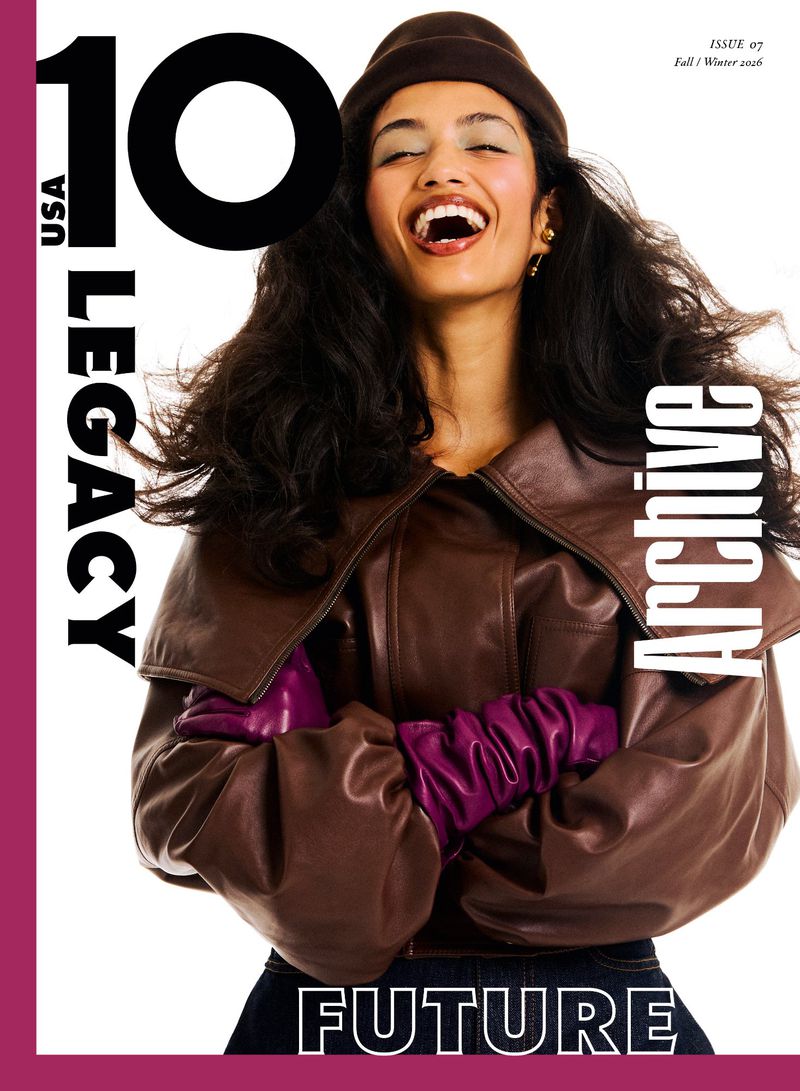 A smiling model wearing an oversized brown leather jacket, purple leather gloves, and a brown beanie on a white studio background.