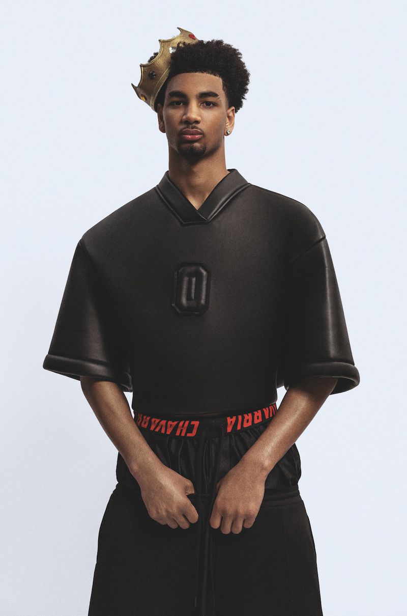 Young adult male in an oversized black jersey and black shorts with red waistband detail posing against a plain white backdrop.