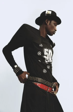 A single Black male model strikes a confident pose in a black t-shirt, hat, and studded belts against a light backdrop in a fashion photo.