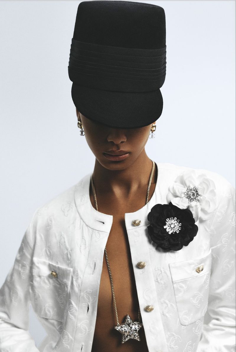 Fashion shot of a woman in a Chanel jacket, hat, and jewelry against a white background, showcasing couture style and elegant design.