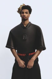 Young black man wearing a crown and a black short-sleeved top against a white studio background.