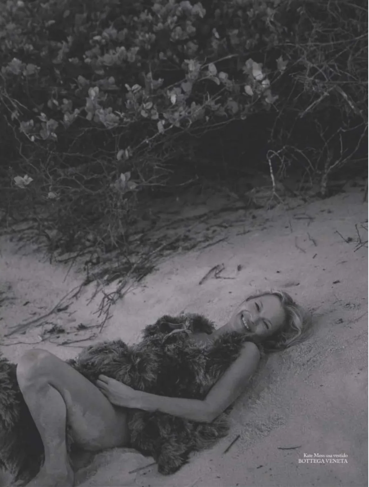Kate Moss smiles while lying on sandy ground beside coastal foliage wearing a textured Bottega Veneta dress in black and white.