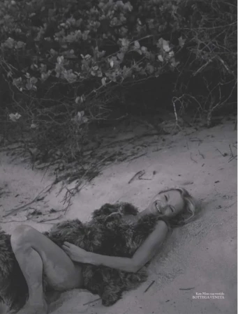 Kate Moss smiles while lying on sandy ground beside coastal foliage wearing a textured Bottega Veneta dress in black and white.