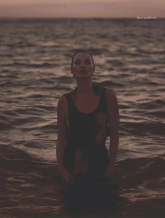 Kate Moss stands waist-deep in the dark ocean water at dusk, wearing a sleeveless black top.