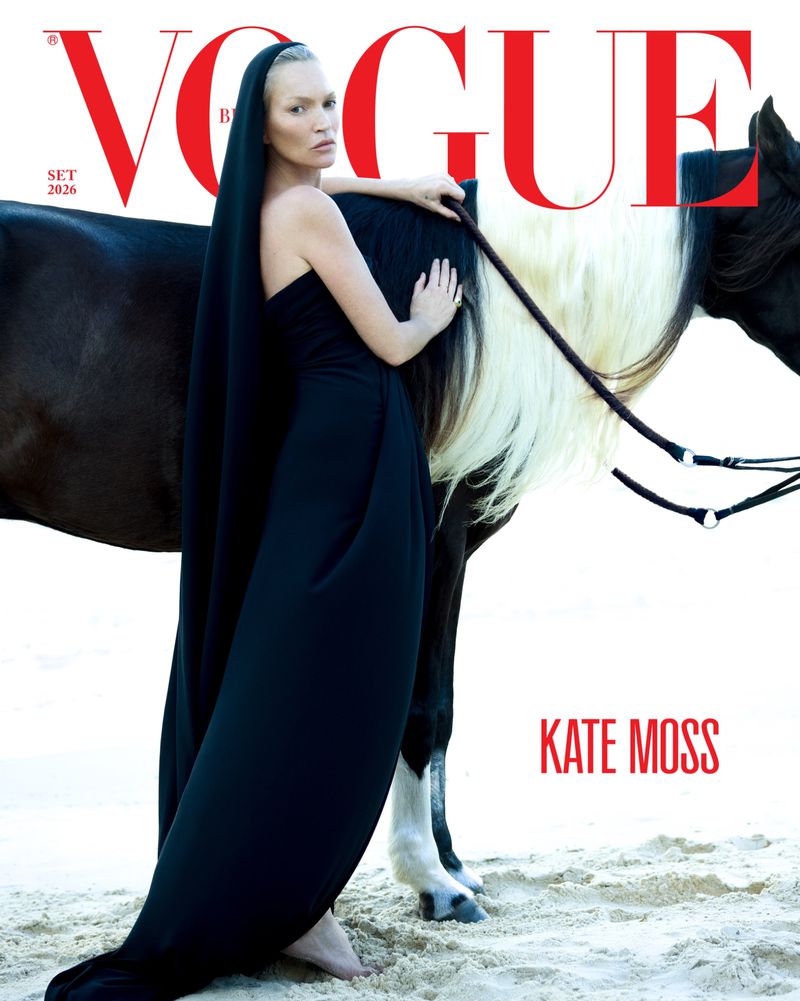 Kate Moss wearing a flowing black hooded gown while leaning against a dark horse with a white mane on a sandy beach.