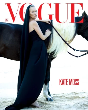Kate Moss wearing a flowing black hooded gown while leaning against a dark horse with a white mane on a sandy beach.