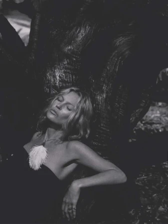 Monochrome portrait of Kate Moss wearing a feather pendant necklace while leaning back against a large tree trunk.