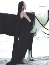 Kate Moss wearing a long black hooded Balenciaga gown standing barefoot beside a black horse on a sandy beach.